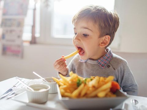 Portrait of small little cute caucasian boy kid eating french fries potato chips at the table in the restaurant or at home three or four years old