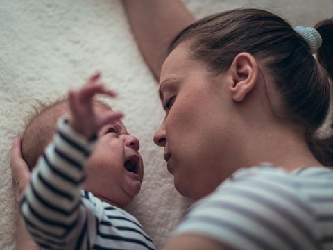 Young single mother holds a baby boy in matching white and blue striped clothes while he is crying. They are lying down on a white blanket.
