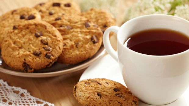 Pile of homemade oatmeal cookies with chocolate chips upon a cutting board on rustic wooden table. Cookies are surrounded by oat flakes, oat spikes and chocolate chips. Main focus is on cookies and in the defocused background is a glass full of milk. Low key DSLR photo taken with Canon EOS 6D Mark II and Canon EF 24-105 mm f/4L