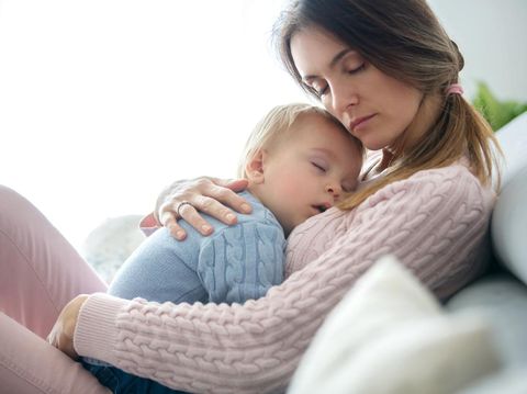 Young mother, holding her sick toddler boy, hugging him at home, sunny living room
