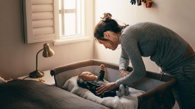 Mother putting her baby to sleep on a bedside baby crib. Woman bending forward over a crib to check her sleeping baby.
