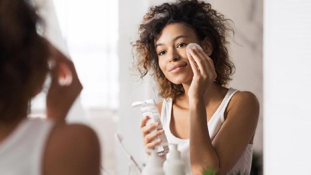 Young afro-american woman cleaning face with cotton pad looking in mirror at bathroom
