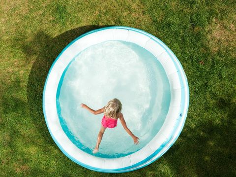 A six year old girl is jumping in an inflatable swimming pool on a hot Summer day. The grass is becoming dry and brown.