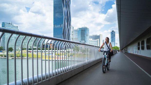 smiling 45 years old businesswoman cycling on her electric bicycle on bridge in office district in Vienna on summer day