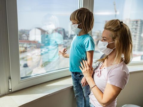 Single mother and her small son with face masks looking through window while being in isolation at home. Focus is on boy.
