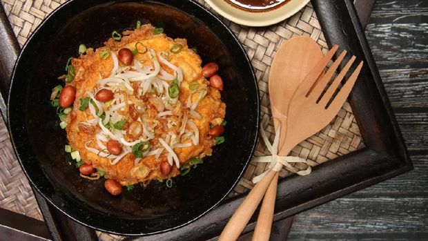 Tahu Telur, a popular egg dish in Surabaya, East Java. Served on black ceramic plate and topped with beansprouts, peanuts, and scallion. Accompanied with spicy soy sauce aside. A pair of wooden cutlery tied with straw ribbon is placed next to the plate. The dish is served on a woven tray stacked on top of another one.