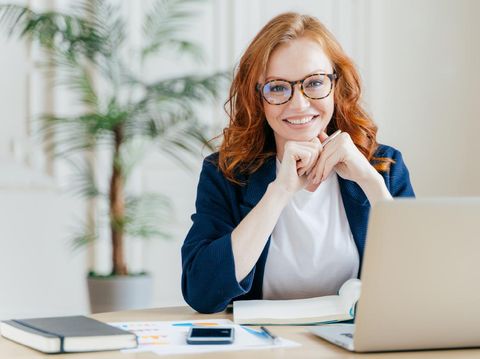 Young Caucasian busy brunette smiling and holding pencil while sitting in office and looking at laptop.