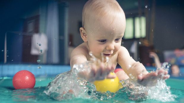 Nine month old baby boy at his first swimming lesson. Smiling, enjoying water and splashing.