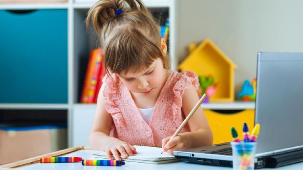 Beautiful little girl writing homework using notebook for distance learning in the kids room. Distance online learning concept.