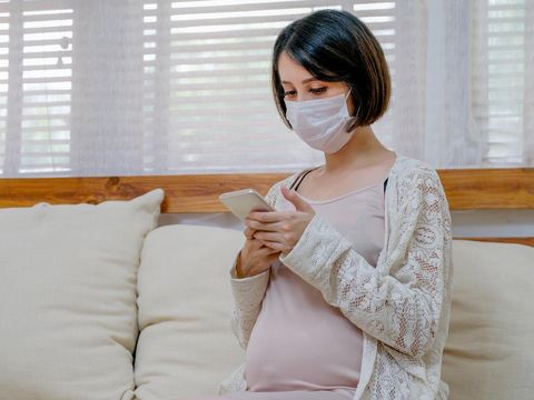 Asian pregnant woman with mask look at mobile phone and sit on sofa in living room of her house with soft light.