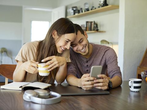Cheerful boyfriend showing mobile phone to girlfriend in dining room. Young woman is holding coffee cup. They are spending leisure time together at home.
