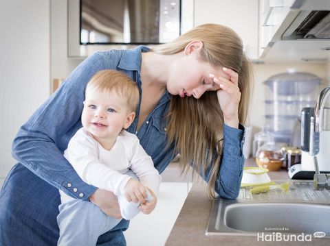 Tired concerned new mother rocking crying baby in kitchen. Portrait of young woman and cute little child in home interior. Motherhood concept
