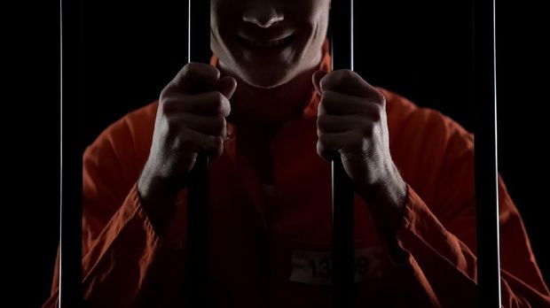 Insane maniac holding prison cell bars waiting for death penalty, smirking