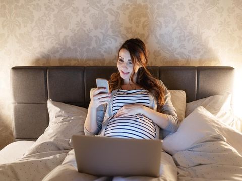 Young smiling pregnant brunette lying in bed with laptop on thighs and using smart phone. Evening time.