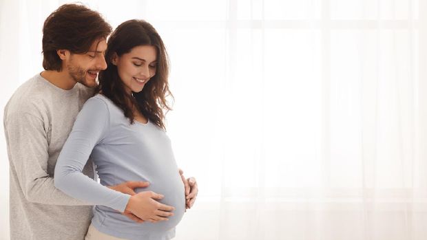 Future parents. Happy expecting couple hugging and smiling while standing near the window at home, free space