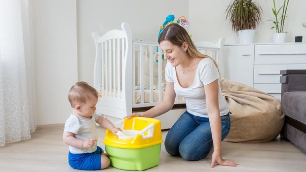 Young smiling mother sitting on floor at living room and teaching her 10 months old baby boy how to use chamber pot