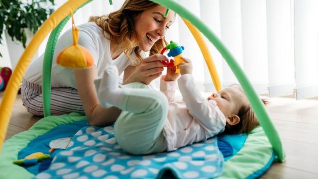 Young smiling woman sitting in her home and playing with her baby. Baby is lying on the floor on baby blanket. They are happy and smiling. Mother holding toy and her baby trying to reach it.