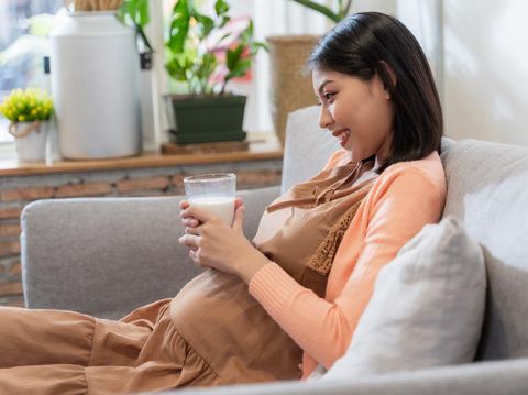 Asian pregnant woman hold a glass of milk for her health and baby with smile and sitting on the sofa, feeling happily and relaxed. Pregnant women concept.