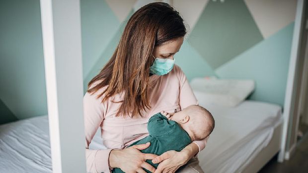 Two people, mother with protective mask breastfeeding her baby son at home, they are at home do to pandemic outbreak quarantine.