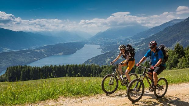 ilustrasi bersepeda A couple is biking uphill on a scenic forest road with an overview of Lake Millstatt, Carinthia, southern Austria. The region is a very attractive holiday area and is famous for hiking, mountainbiking and swimming during summertime. In wintertime there lots of skiing areas as well.