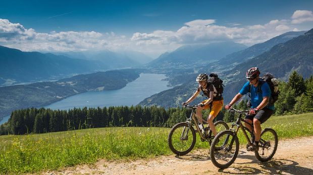 A couple is biking uphill on a scenic forest road with an overview of Lake Millstatt, Carinthia, southern Austria. The region is a very attractive holiday area and is famous for hiking, mountainbiking and swimming during summertime. In wintertime there lots of skiing areas as well.