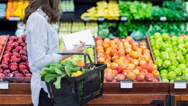 Young woman carries a shopping basket filled with fresh produce. She is shopping for fresh fruit and vegetables in a grocery store.