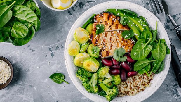 Healthy buddha bowl lunch with grilled chicken, quinoa, spinach, avocado, brussels sprouts, broccoli, red beans with sesame seeds on dark gray background. Top view.