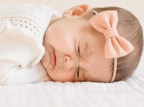 A 6-month old baby girl wearing a white outfit and a peach-colored bow laying on a white blanket, sneezing.