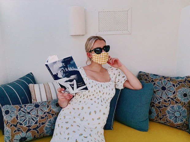 Terlihat Emma mengenakan floral sundress puff sleves, dengan kacamata hitam, masker motif kotak-kotak sambil memegang buku