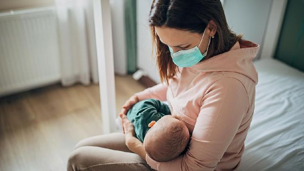 Two people, mother with protective mask breastfeeding her baby son at home, they are at home do to pandemic outbreak quarantine.