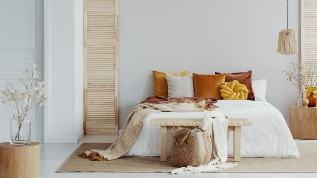Brown and orange pillows on white bed in natural bedroom interior with wicker lamp and wooden bedside table with vase