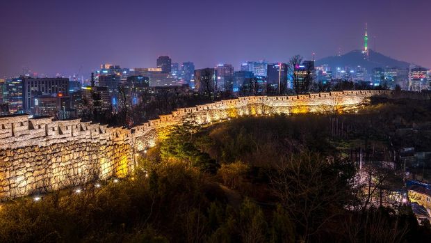 The old city wall lit up in evening as the lights of downtown Seoul shine in the distance.