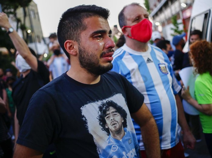 A man wearing a shirt with the face of Diego Maradona cries outside the stadium of the Argentinos Juniors soccer club, where he started as a professional footballer, in Buenos Aires, Argentina, Wednesday, Nov. 25, 2020. The Argentine soccer great who was among the best players ever and who led his country to the 1986 World Cup title died from a heart attack at his home in Buenos Aires. He was 60. (AP Photo/Marcos Brindicci)