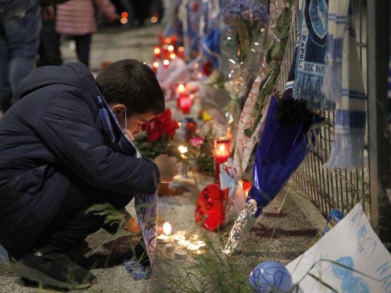 A boy lights a candle as people gather outside the San Paolo Stadium to pay their homage to soccer legend Diego Maradona, in Naples, Italy, Wednesday, Nov. 25, 2020. Diego Maradona has died. The Argentine soccer great was among the best players ever and who led his country to the 1986 World Cup title before later struggling with cocaine use and obesity. He was 60. (AP Photo/Alessandra Tarantino)