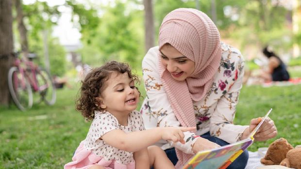 A young Arab American mother reads with her young daughter in a public park. She is wearing a hijab and having a great time as they turn pages and point to pictures together.
