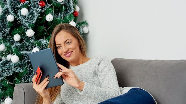 Caucasian woman lying down on sofa next to Christmas tree and using tablet for on-line shopping