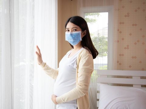 Young beautiful pregnant asian woman standing and touching belly near the window in bedroom. Pregnancy, motherhood wearing virus protection mask mouth concept.