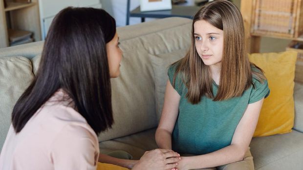Upset teenage girl sitting on sofa and sharing sufferings with mother holding her hands