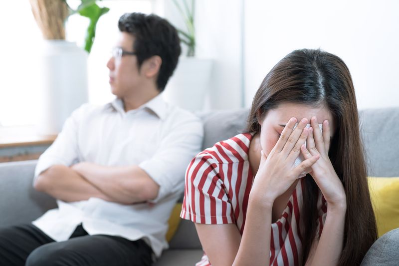 An Asian wife is crying on the sofa after an argument with her husband behind her.