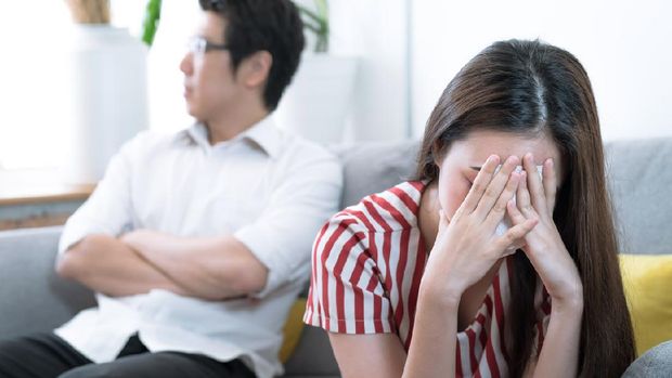 An Asian wife is crying on the sofa after an argument with her husband behind her.