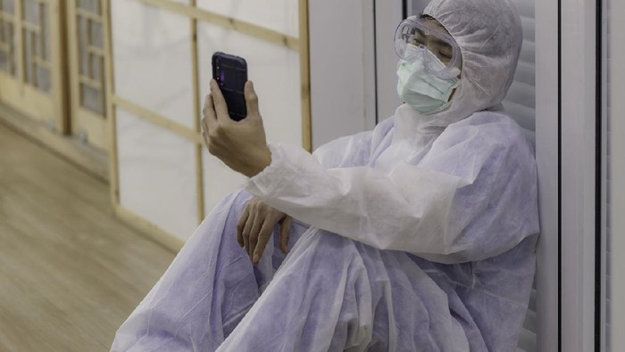young Asian doctor, dressed in anti-virus clothing, sits on the floor tired and uses a smartphone to make a video call to his family. corona virus concept.