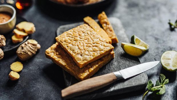 Close-up of tempeh on cutting board with kitchen knife with other ingredients on table. Preparing a vegan dish in kitchen.