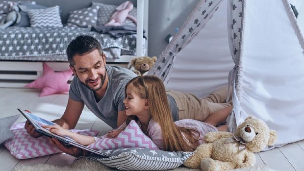 Father reading a book to his daughter while lying on the floor in bedroom