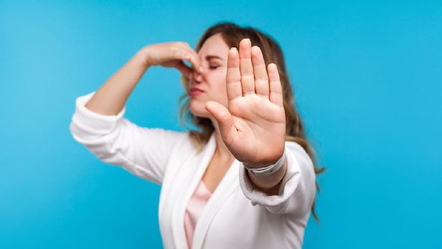Bad smell. Portrait of woman with wavy hair in white jacket pinching her nose and showing stop gesture, expressing disgust to unpleasant odor, fart gases, her eyes closed with abhor. blue background