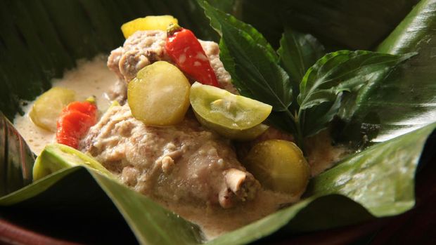 Garang Asem Ayam, the traditional Javanese tangy chicken curry. Each portion is placed in a banana leaf pouch then steamed. One portion is served and plated on an earthenware plate. Some fresh green tomatoes, bilimbi fruits, and unopened pouches are arranged on the table.