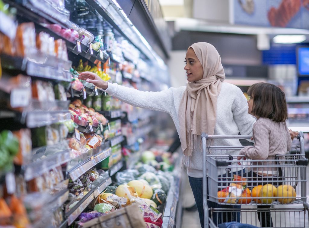 A young Muslim mother and daughter walk the produce aisles of the grocery store together shopping for groceries.  They are both dressed casually and the mother is wearing a Hijab.  The mother is reaching for products on the shelf while pushing a shopping cart with her daughter seated in it.