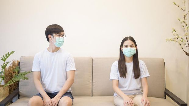 A couple wearing a mask is sitting on a sofa at home during quarantine for protection and avoiding from coronavirus