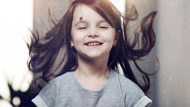 front view of little girl with brown flying hair having toothy smile on her face and feeling joyful, photo taken indoors.