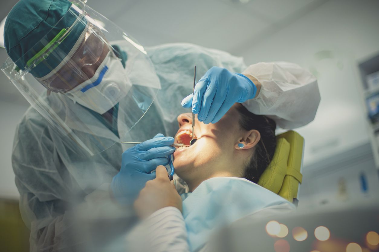 The dentist, fully dressed in a protective suit with an eyewear protection is with a patient. A woman in the chair has her mouth opened and examined while holding a suction pump.
