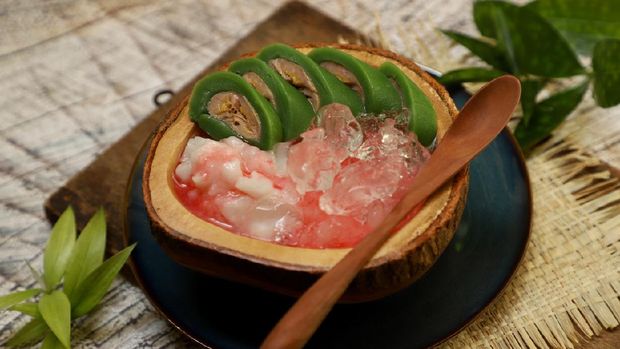 Es Pisang Ijo, the popular dessert of plantain banana roll with coconut milk custard and red sugar syrup from Makassar (Ujung Pandang), South Sulawesi. The banana is wrapped with pandan crêpe; then served in slices on the custard and drenched with red sugar syrup. The dessert is plated on a wooden bowl and placed on ceramic plate. A wooden spoon is placed on the rim of the bowl. Several sprigs of bamboo leaves are used for table decoration.